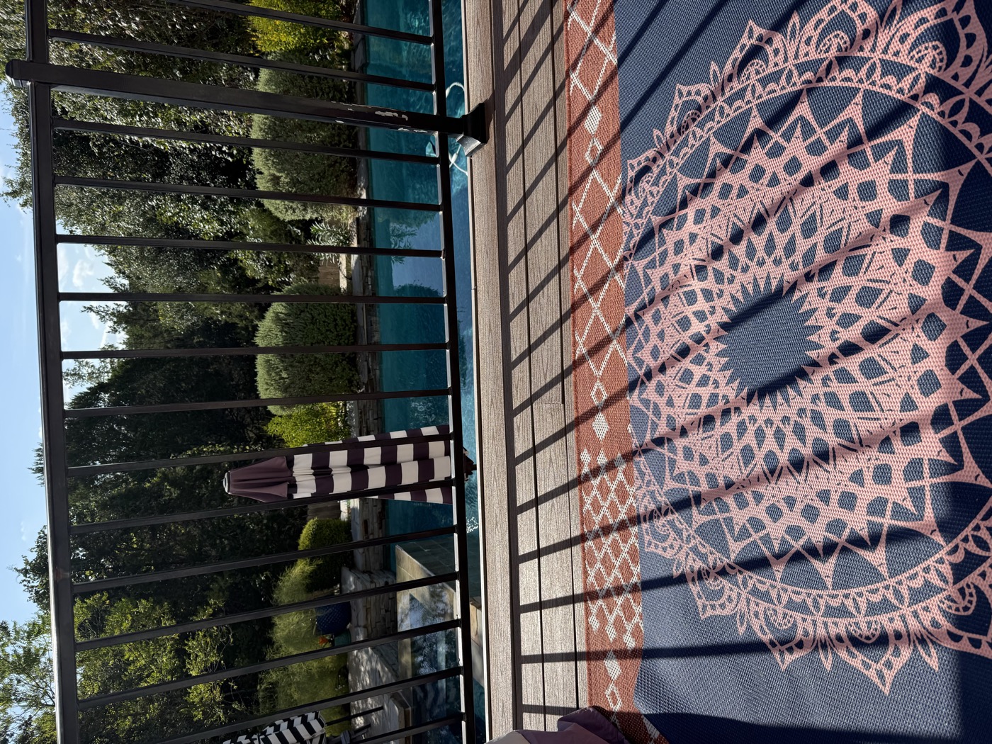 A navy and blush mandala yoga mat on a wooden back deck, with a pool and a wall of green trees beyond the railing in morning sun.