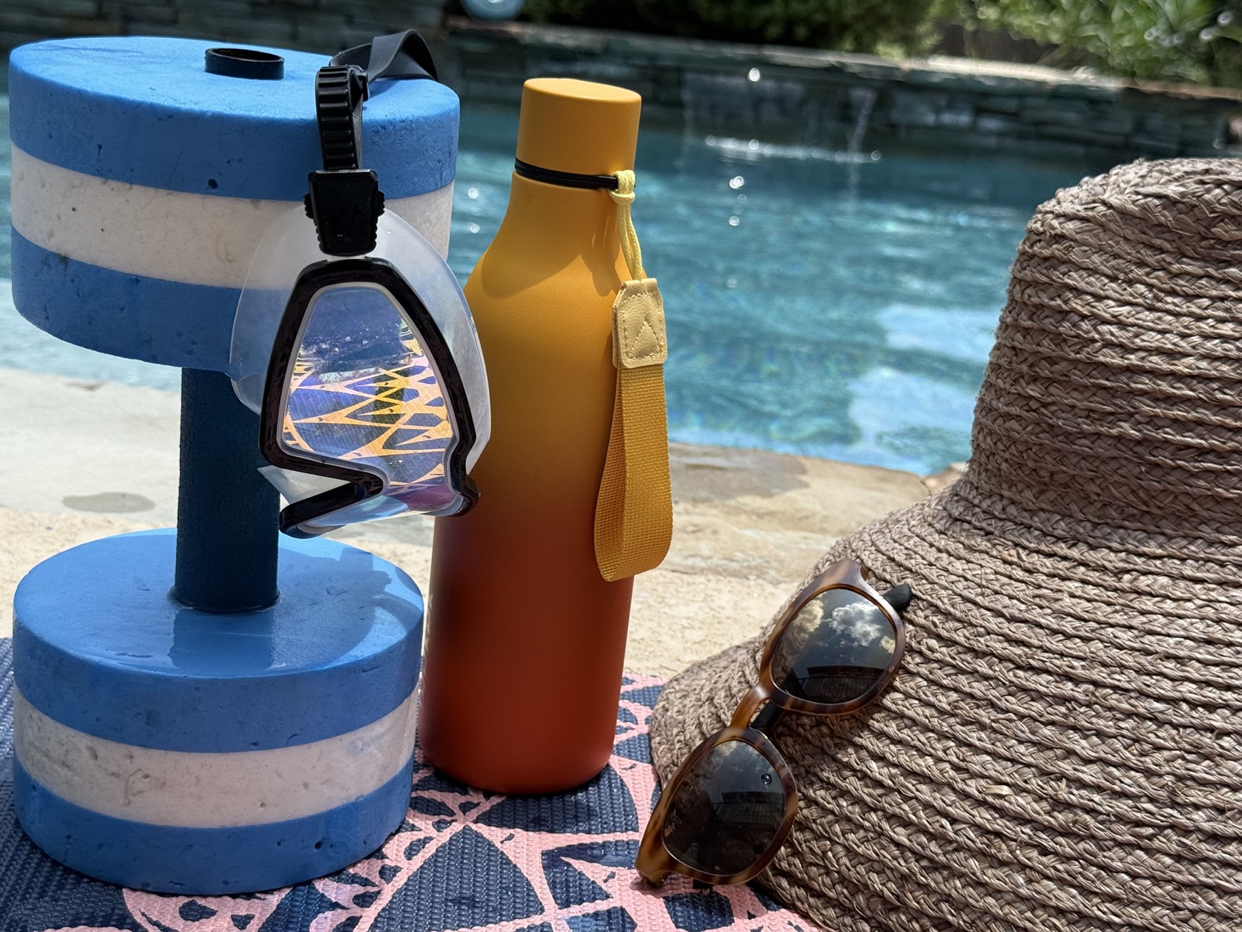 Poolside flat-lay: blue foam water dumbbells, swim goggles, a water bottle, a straw sun hat, and sunglasses.
