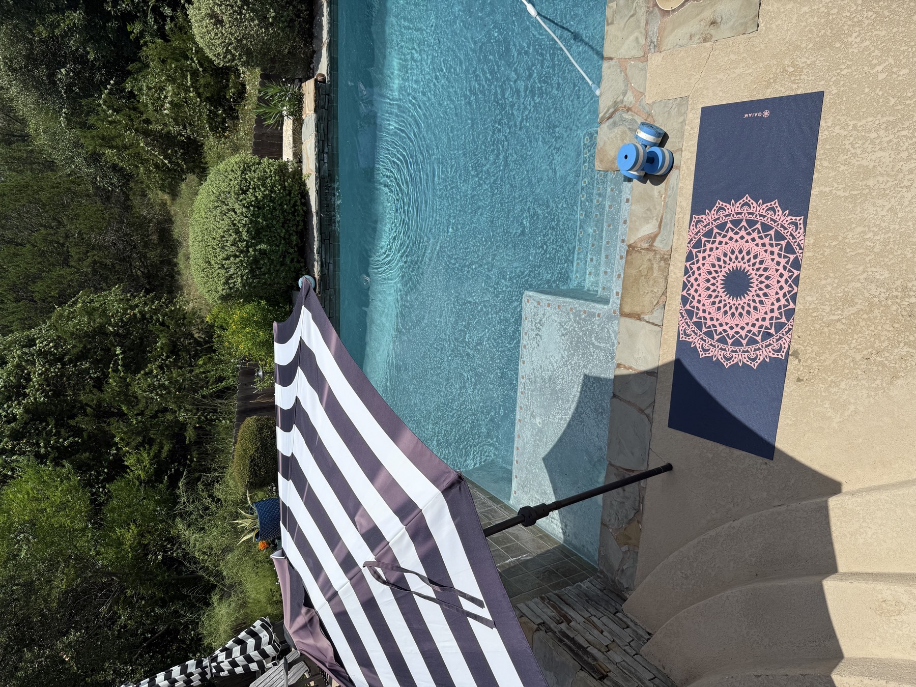 Overhead view of a backyard pool with a striped umbrella, a yoga mat, and blue water dumbbells set out poolside.