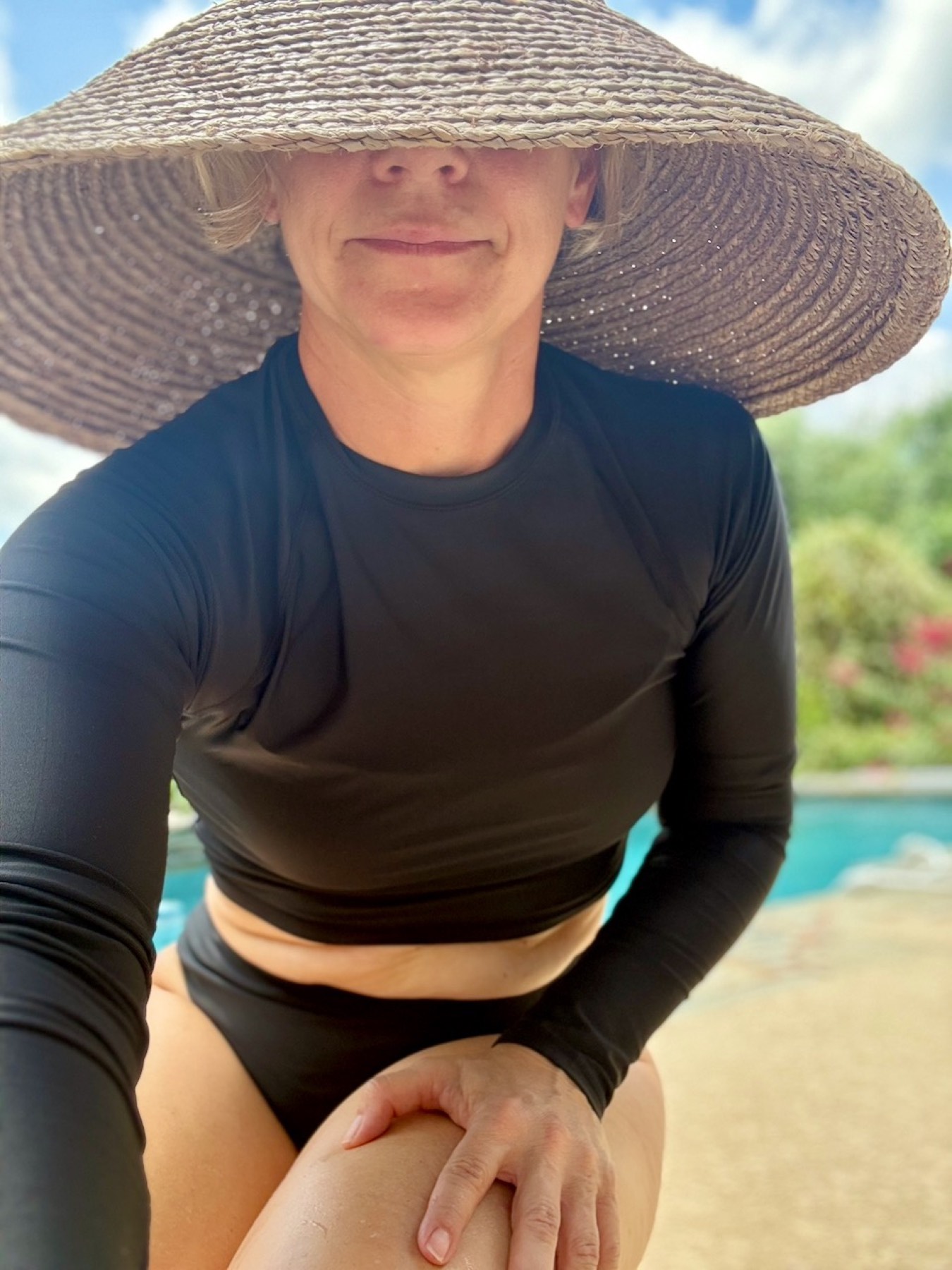 Poolside selfie in a wide-brim straw hat and long-sleeve swim top, smiling.